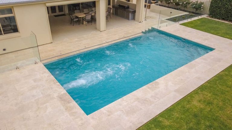 Aerial view of a finished pool with travertine-look paving, frameless glass fencing and a covered outdoor kitchen