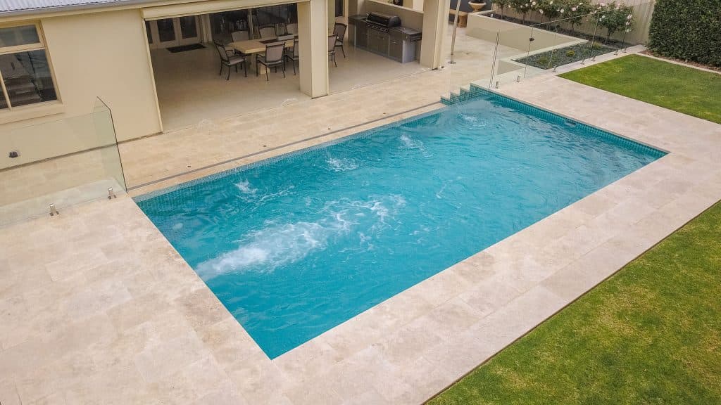 Aerial view of a finished pool with travertine-look paving, frameless glass fencing and a covered outdoor kitchen